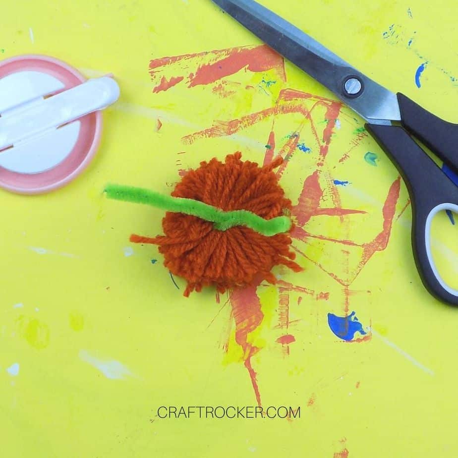 Green Pipe Cleaner Wrapped Around Yarn Pom Pom next to Scissors - Craft Rocker